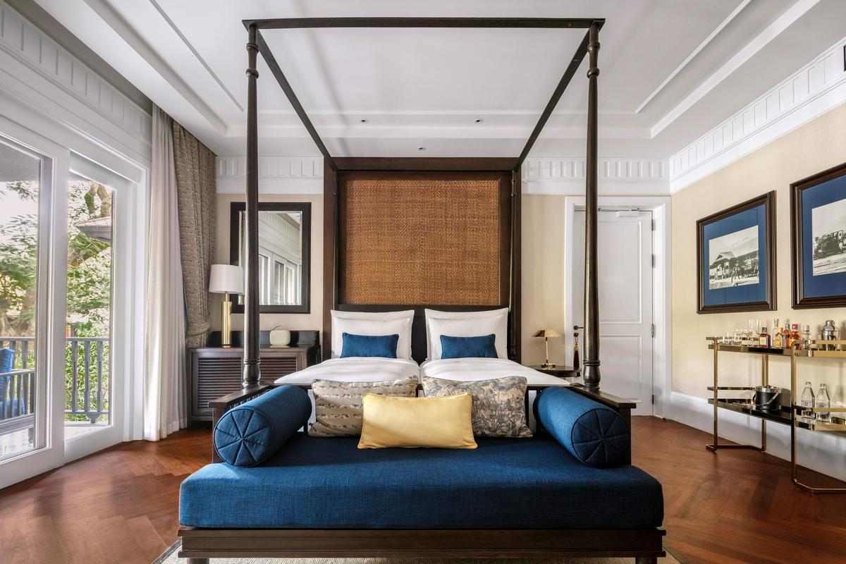 A bedroom of a suite at 137 Pillars Chiang Mai with a dark wood four-poster bed, blue daybed and a gold bar cart against a neutral wall.