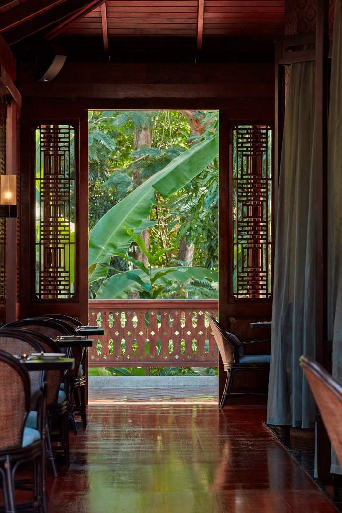 Interior of a dark wood dining room looking out toward bright green tropical leaves through a carved door frame.