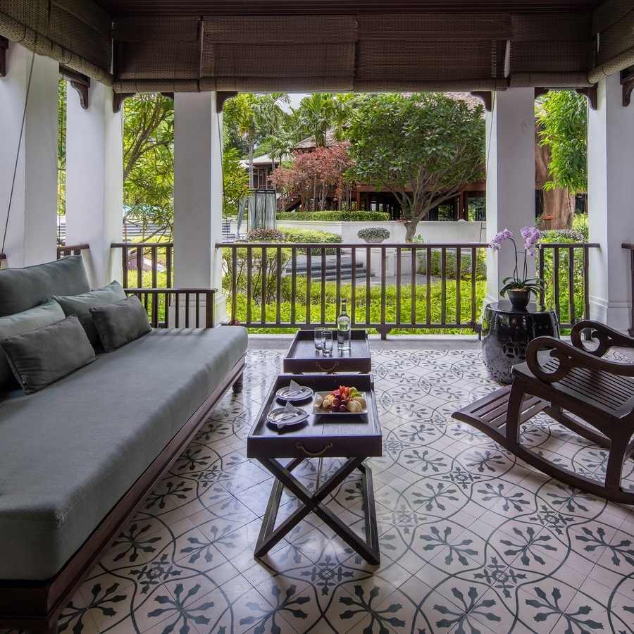 Patterned tile balcony with a gray daybed, wooden rocking chair and outdoor snack trays facing a green garden.
