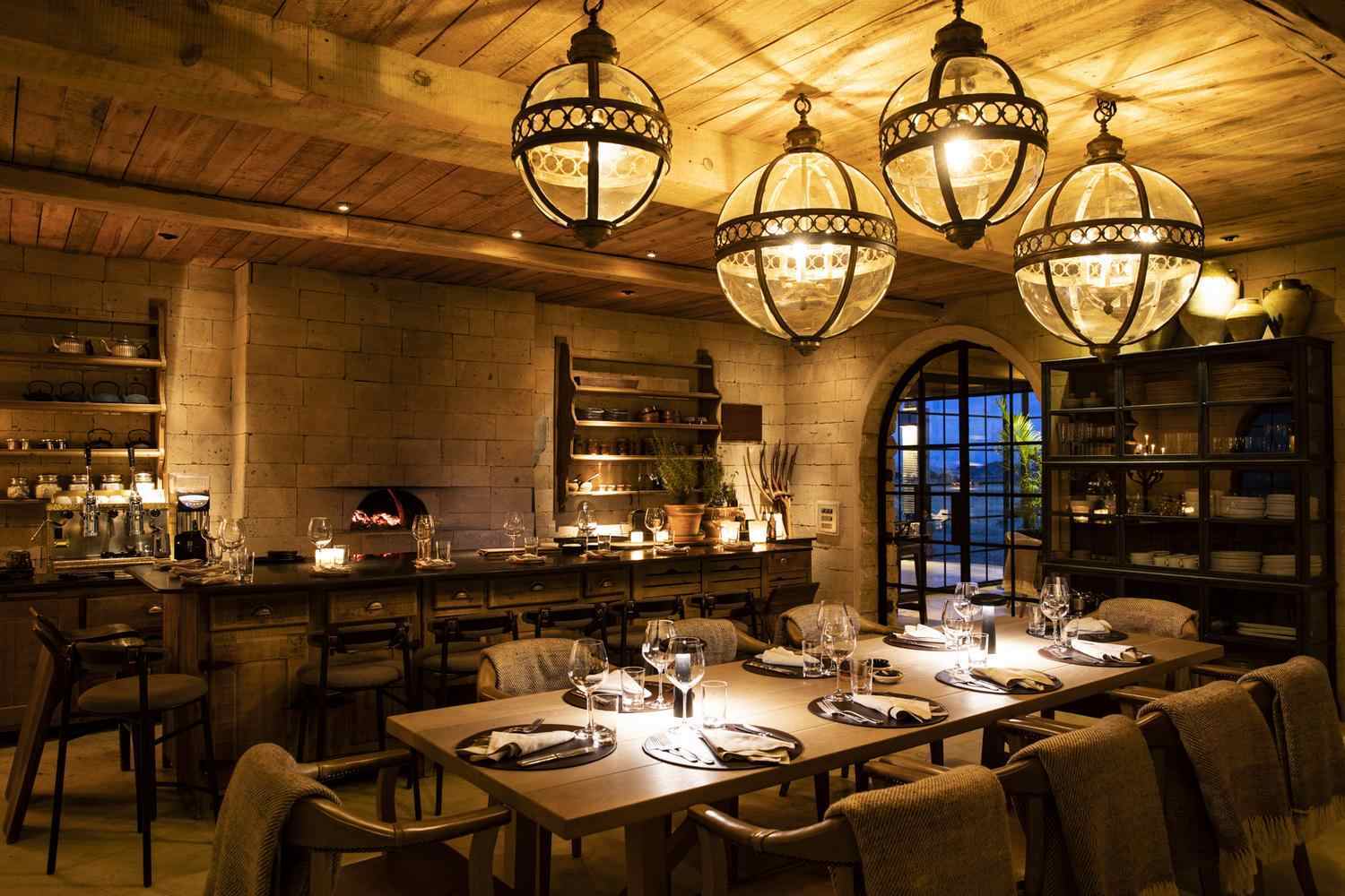 Rustic dining table set with wine glasses under large hanging globe lanterns in a stone-walled room.