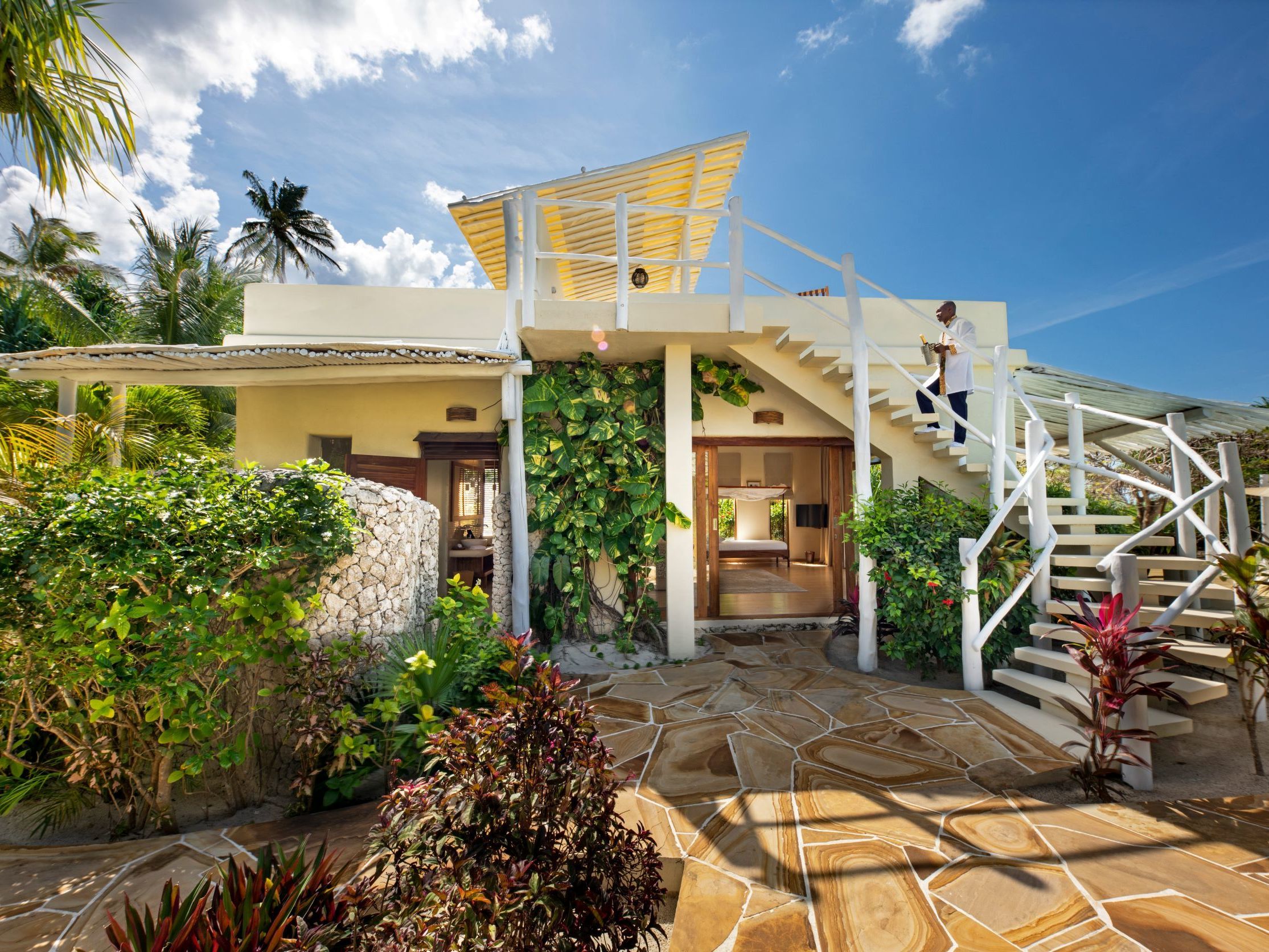 Tropical villa with white staircase and lush garden under a blue sky.