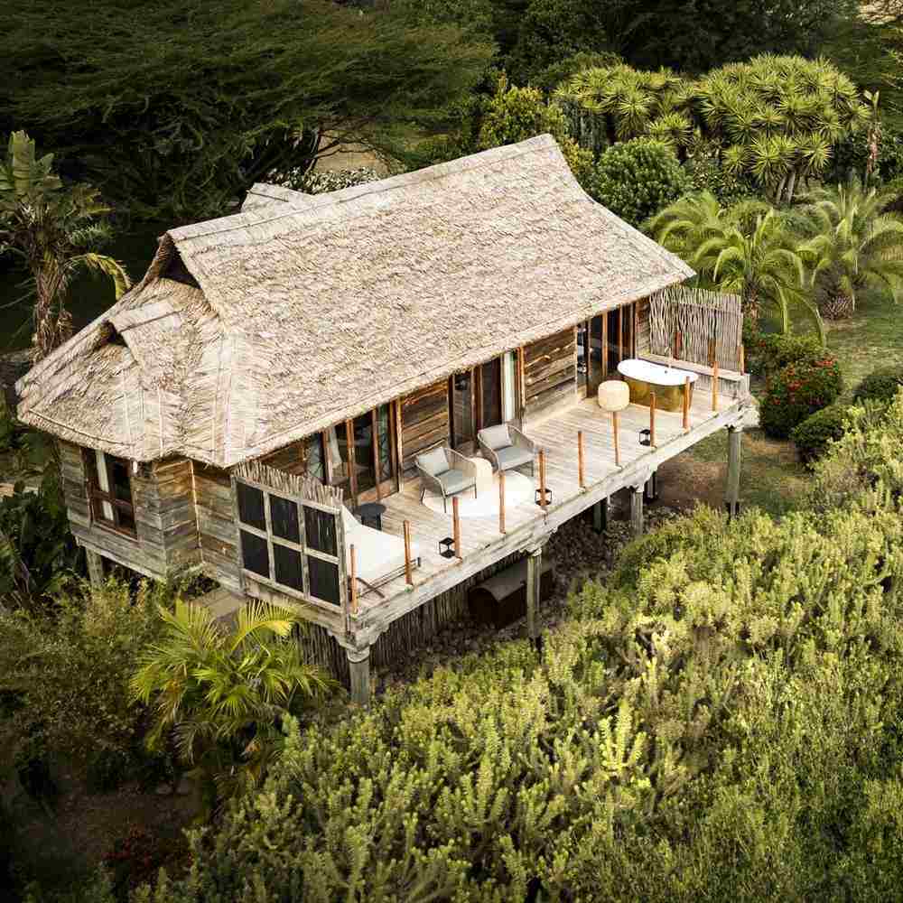 Aerial view of a wooden villa with a thatched roof, balcony seating, and an outdoor bathtub amidst trees.