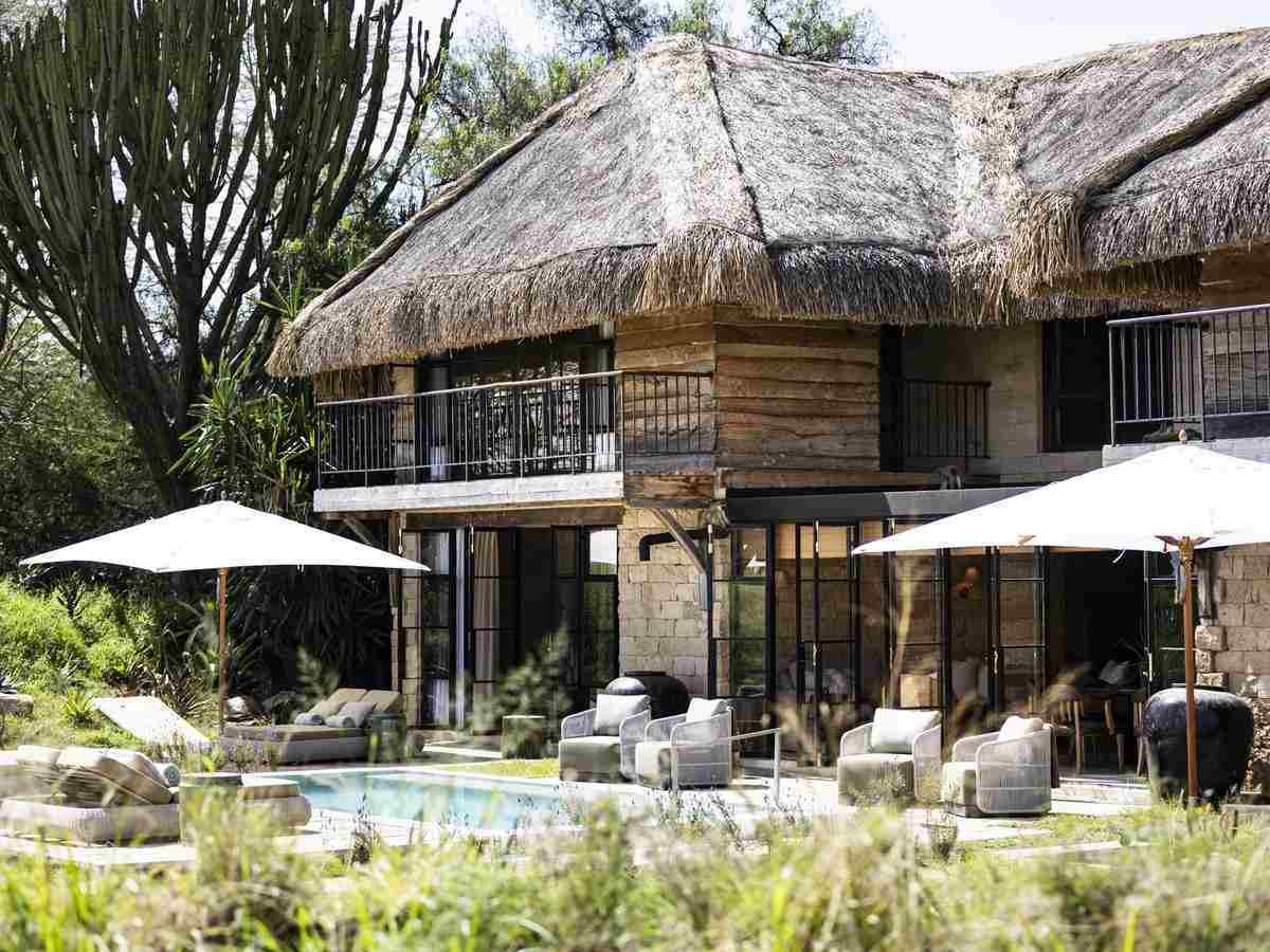Thatched-roof lodge with glass doors opening onto a outdoor patio with lounge chairs, umbrellas, and a pool.