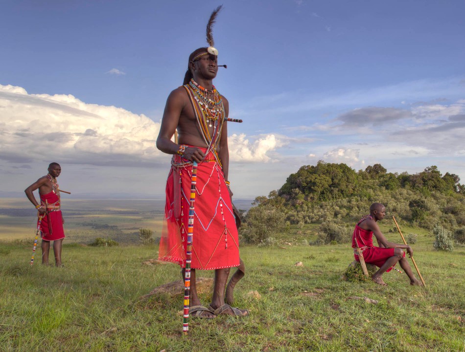 Three men in traditional Maasai attire standing on a green hill against a blue sky with soft white clouds.
