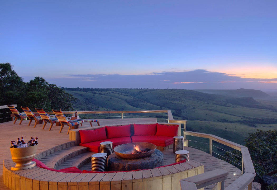 A sunken fire pit with red cushions on a wooden deck overlooking a wide valley under a blue dusk sky.