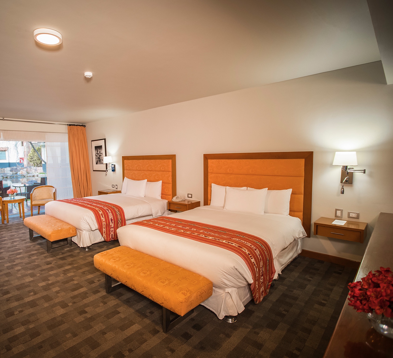 A well-lit hotel room with two double beds, matching orange bedspreads, and a window with curtains.
