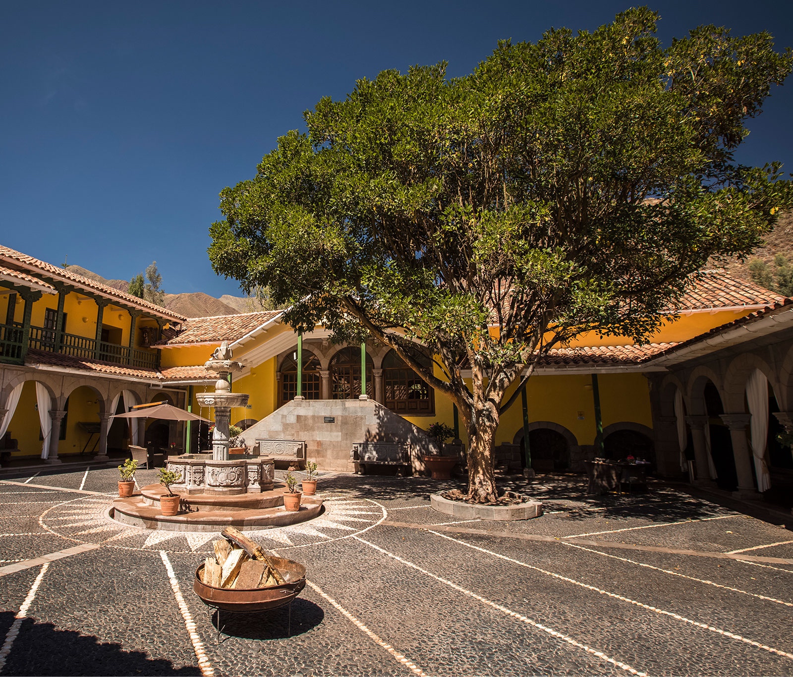 Courtyard with a fountain, large tree, and cobblestone patterns, surrounded by yellow colonial buildings.