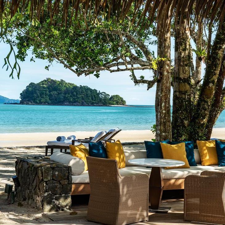 Shaded beach lounge with sofas and wicker chairs looking out at a tropical island in the sea.