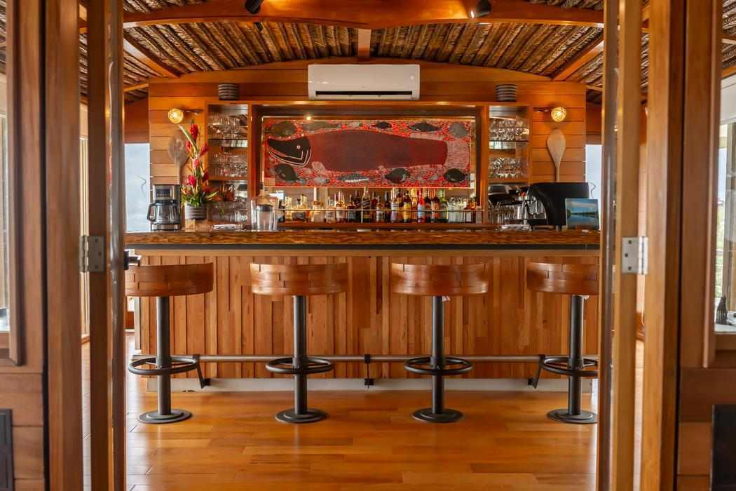 A wooden bar with four stools and shelves of bottles under a thatched ceiling on Delfin I.