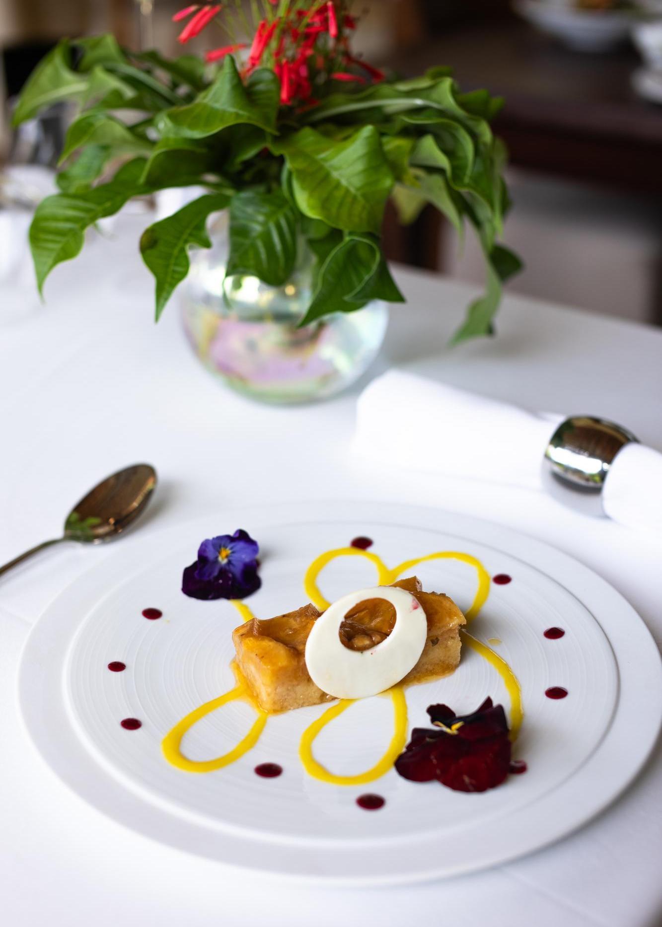 Elegant dessert plating with a pastry, cream, nut topping, and floral garnish on a white plate.