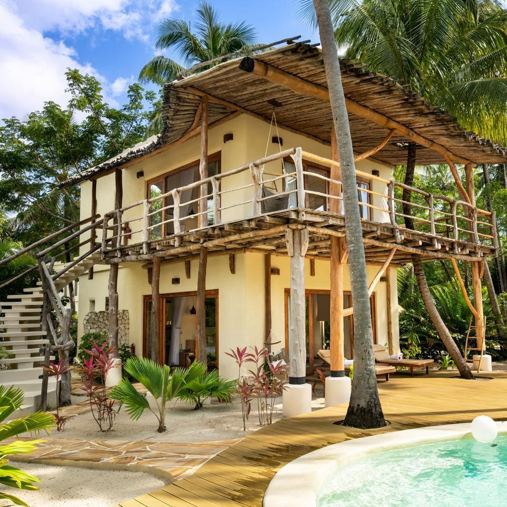 Tropical villa with thatched roof, wooden balcony, and a pool surrounded by palm trees.