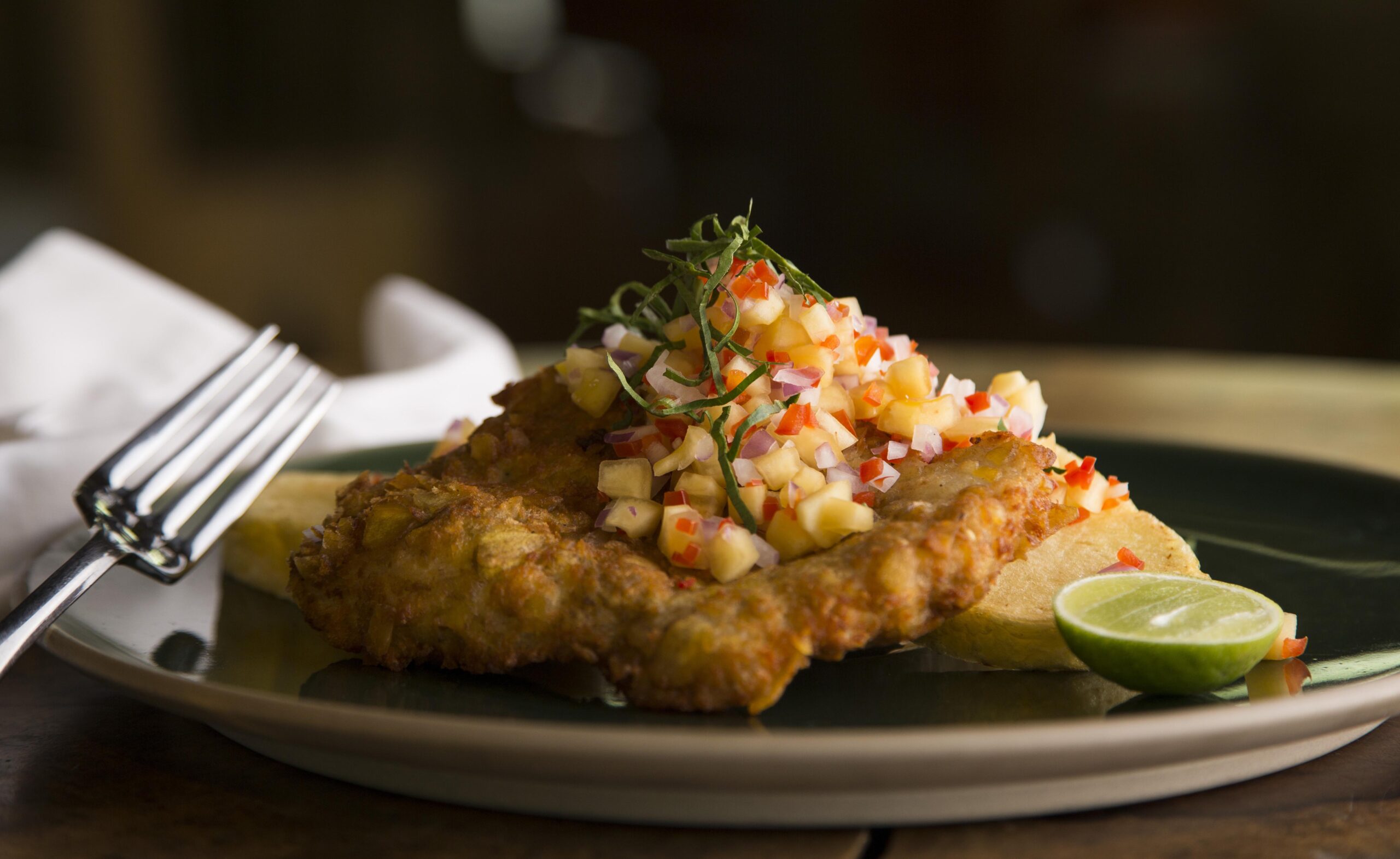 A breaded chicken cutlet topped with diced fruit salsa, served with lime and garnish.