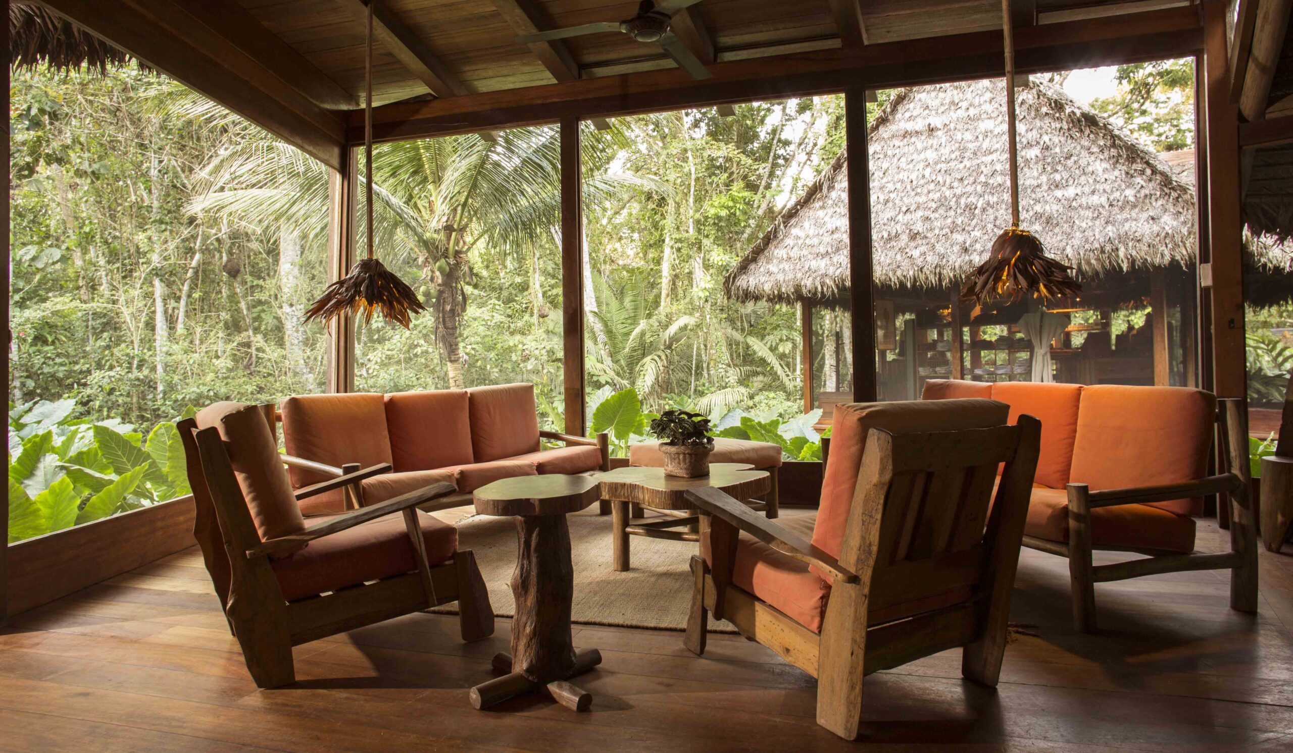 Rustic lounge with wooden furniture and tropical forest view outside large windows.
