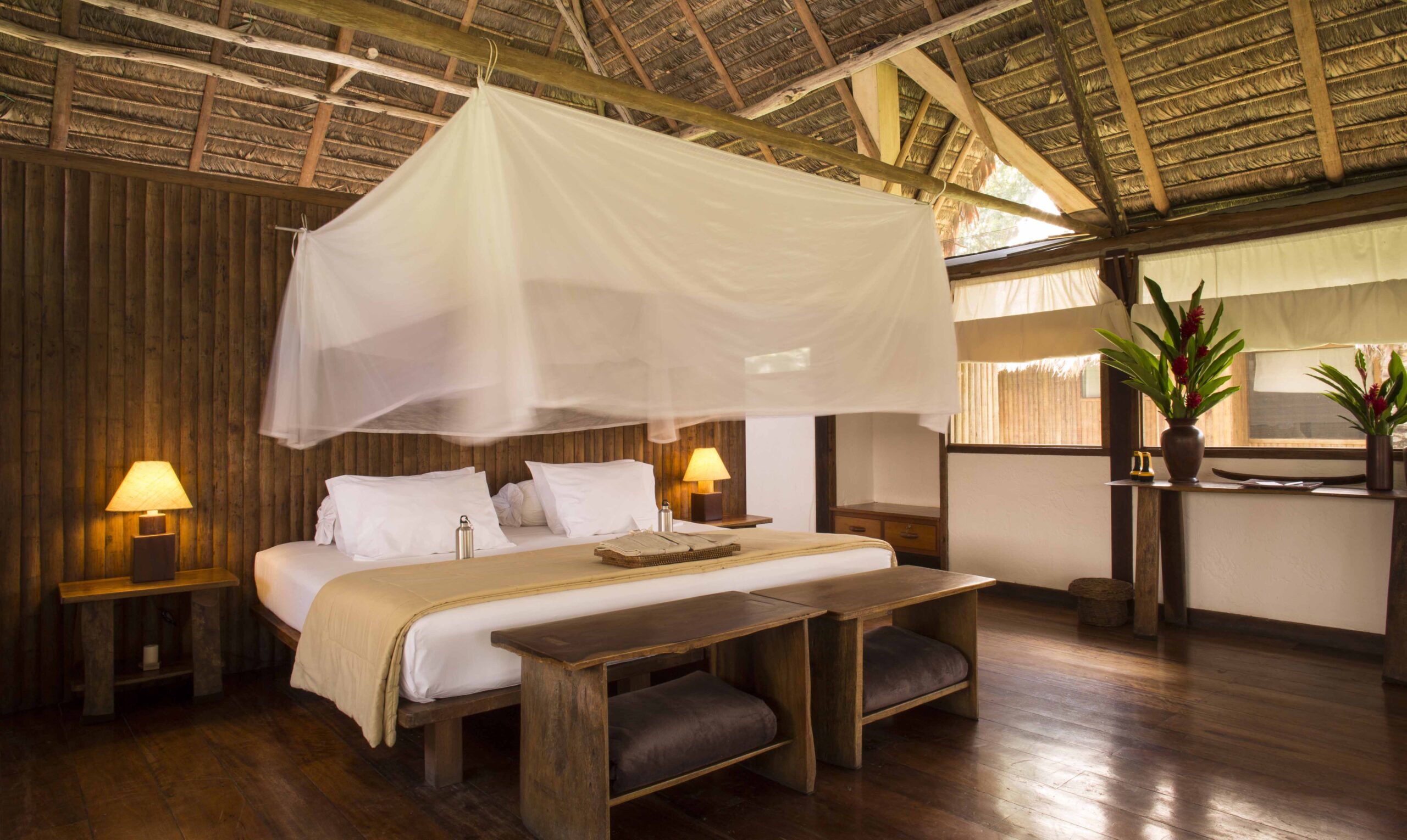 Rustic bedroom with a canopy bed, wooden floors and furniture, and tropical flower arrangements.