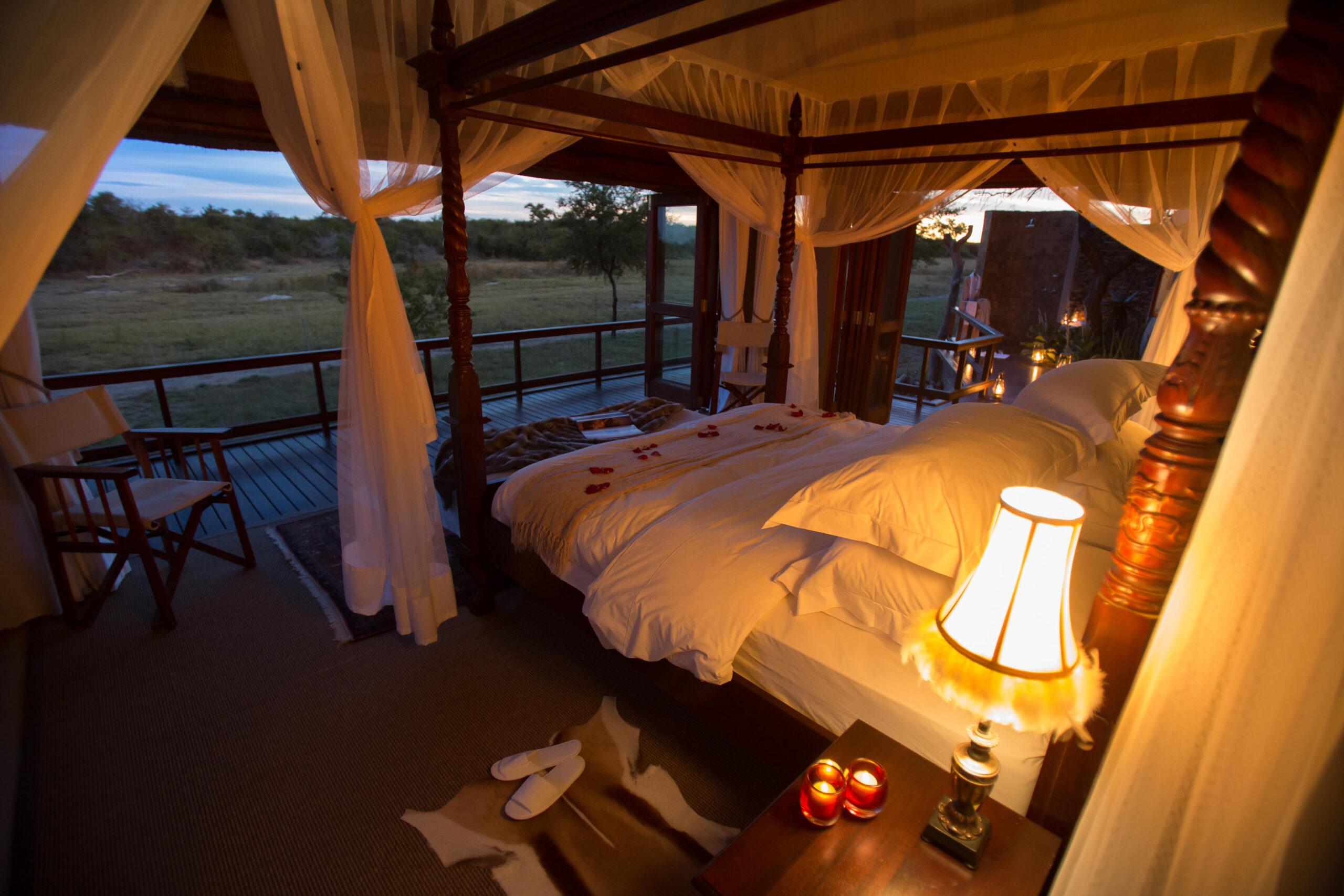 Luxurious safari tent bedroom with canopy bed, warm lighting, and a view of the wilderness at dusk.