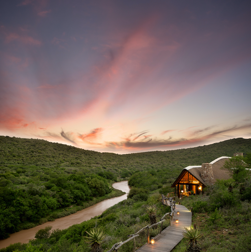 Lodge with lit path at dusk overlooking a river in a lush valley under a vibrant sky.