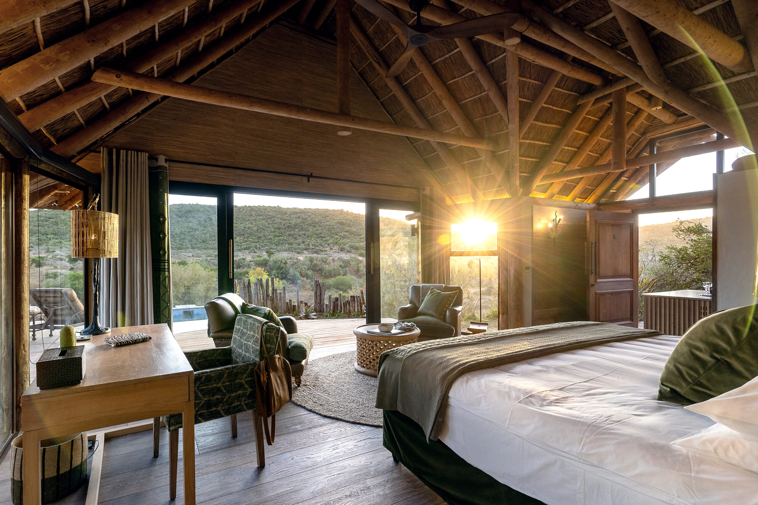A luxurious bedroom with a thatched roof, sliding glass doors opening to hills, and the sun setting.