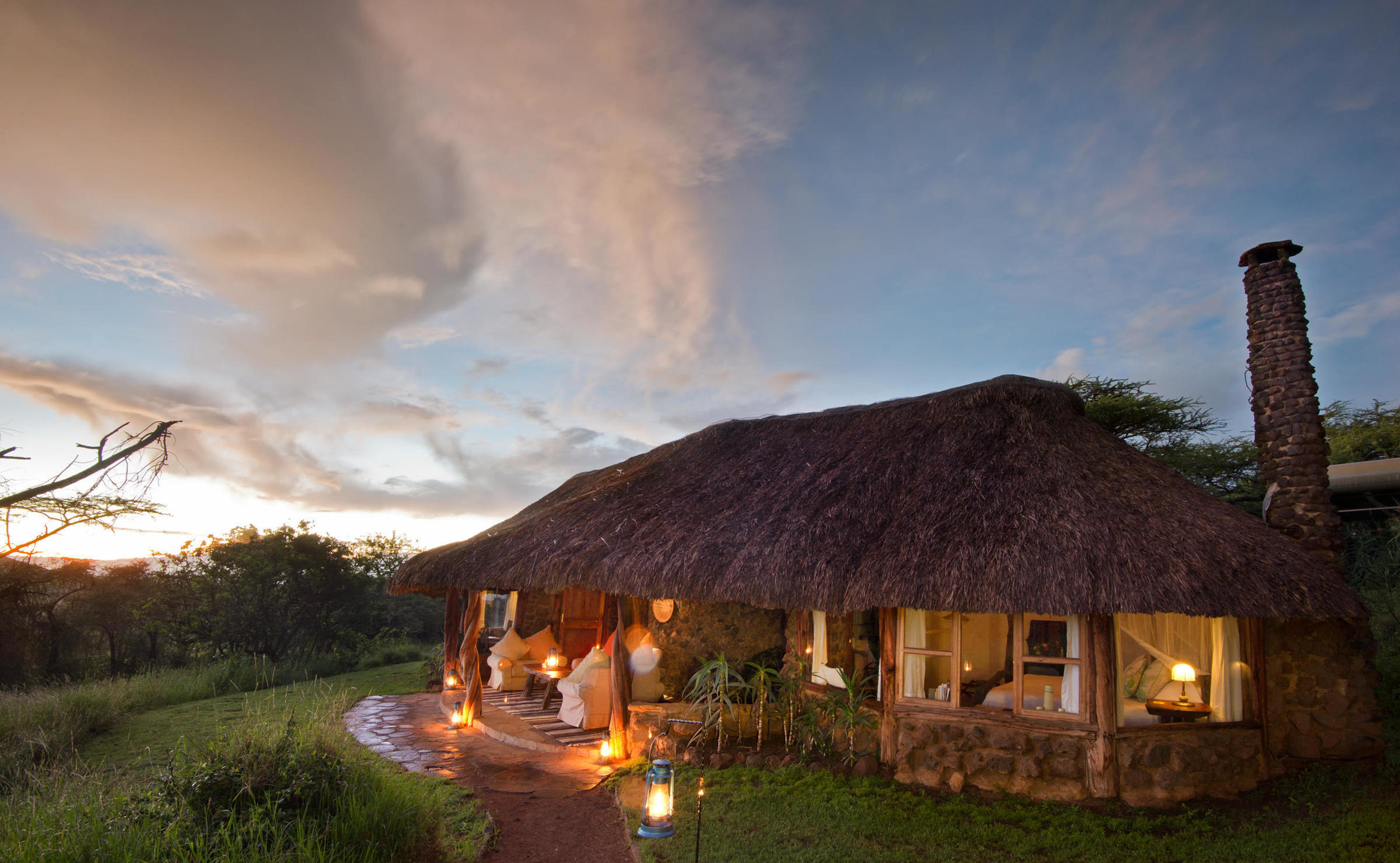 A lit-up thatched cottage at dusk with a stone chimney and lanterns on the porch.
