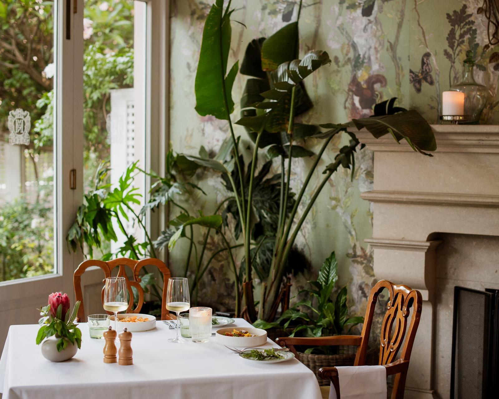 A restaurant table set with white cloth and wine glasses next to a tall tropical plant and a stone fireplace.