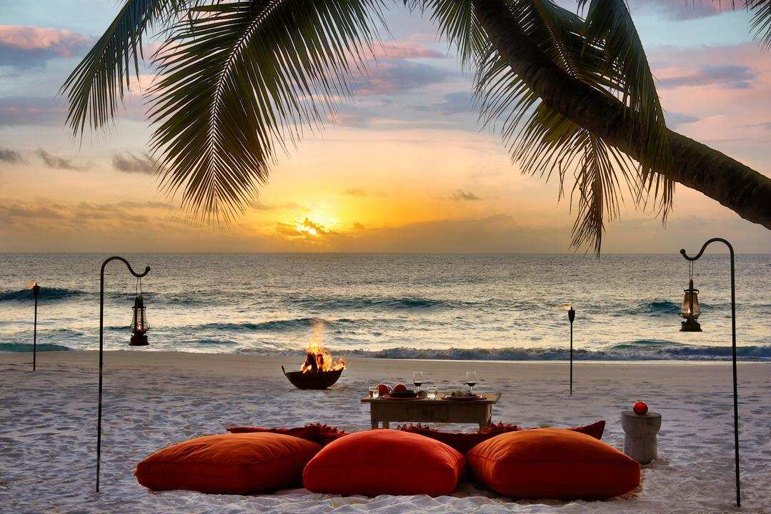 A private beach dinner setup with orange cushions and lanterns on the sand during a vibrant sunset.