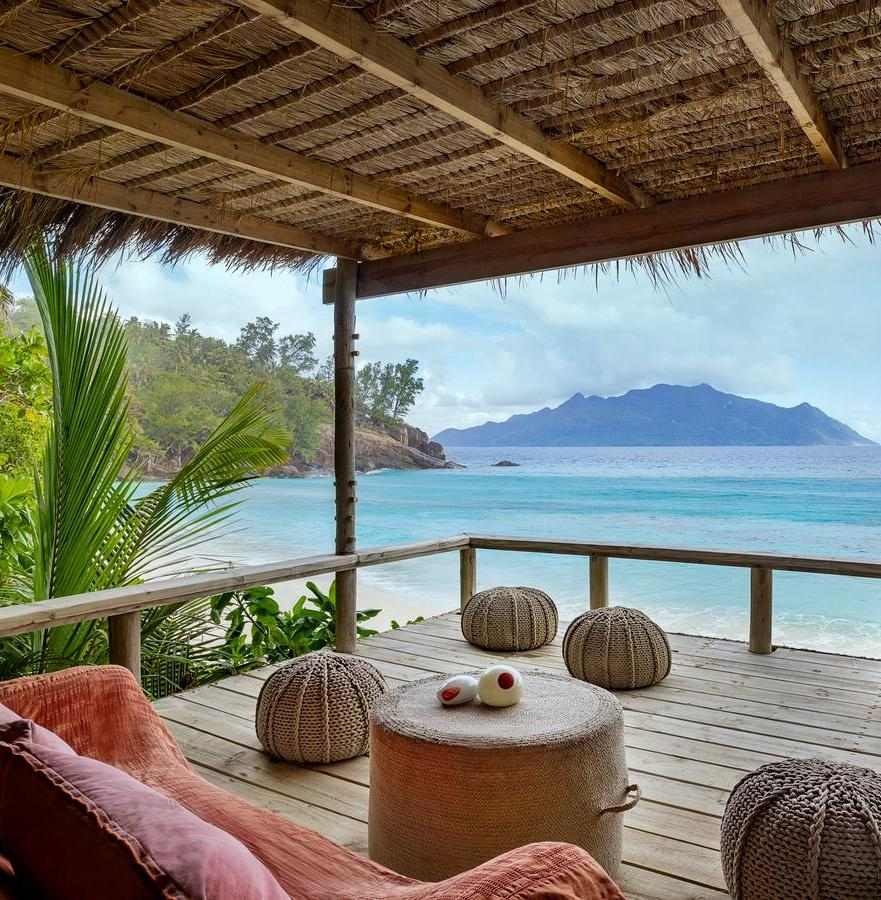 A shaded wooden balcony with outdoor seating and footstools facing a blue ocean and distant island.