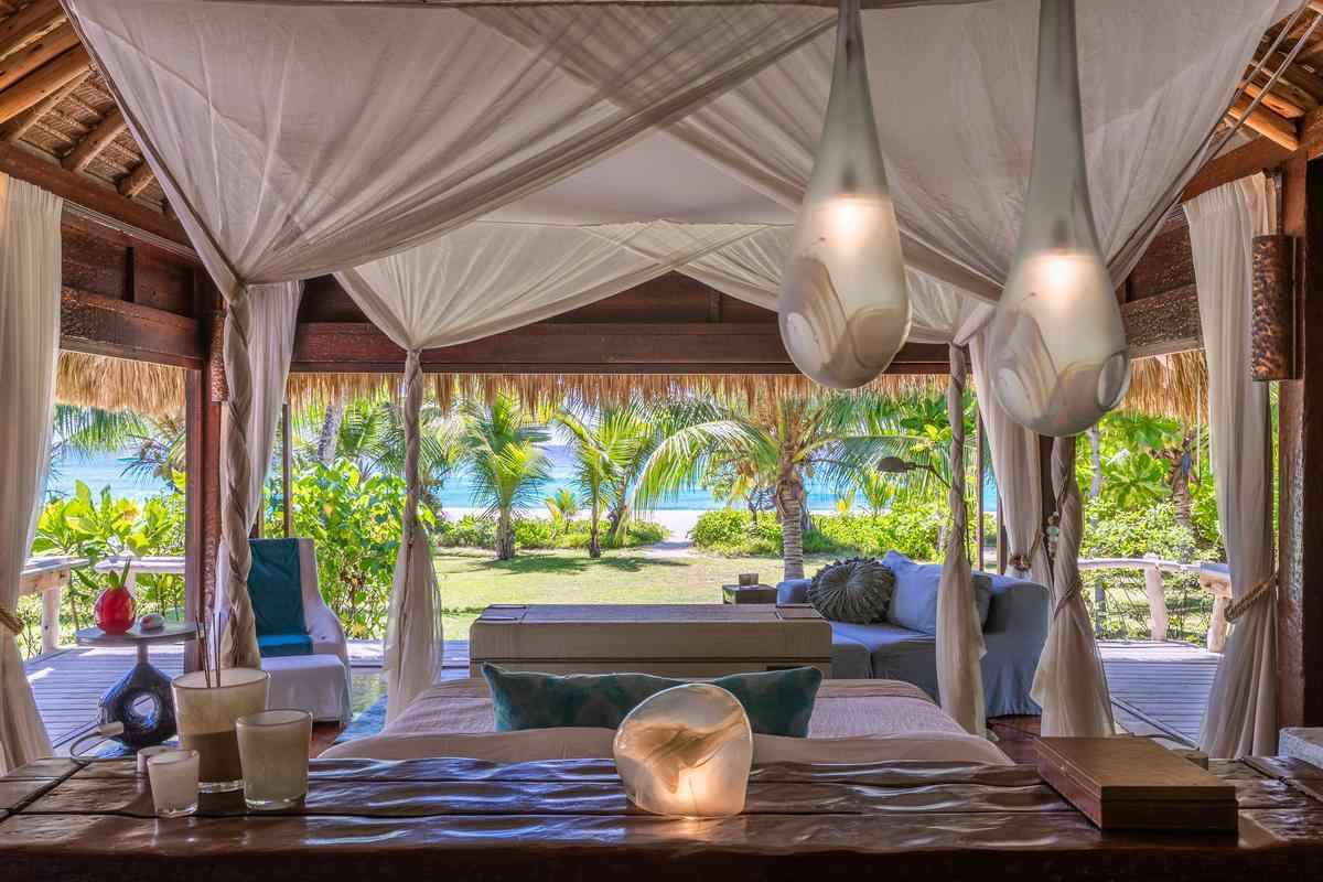 Interior of a luxury bedroom with a four-poster bed looking out toward a tropical beach.