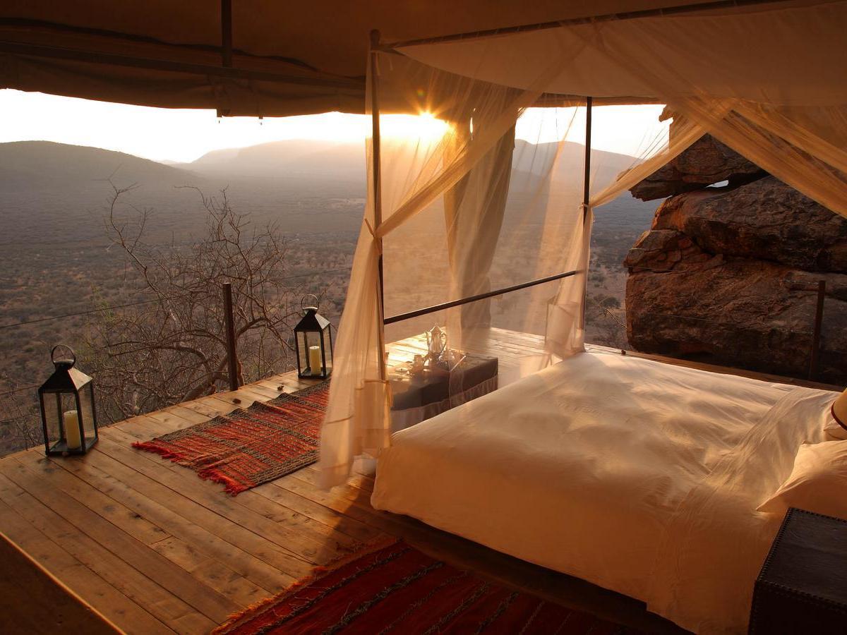 An open-air bedroom suite with a mosquito net bed and lanterns on a wooden deck at sunset in Saruni Samburu.
