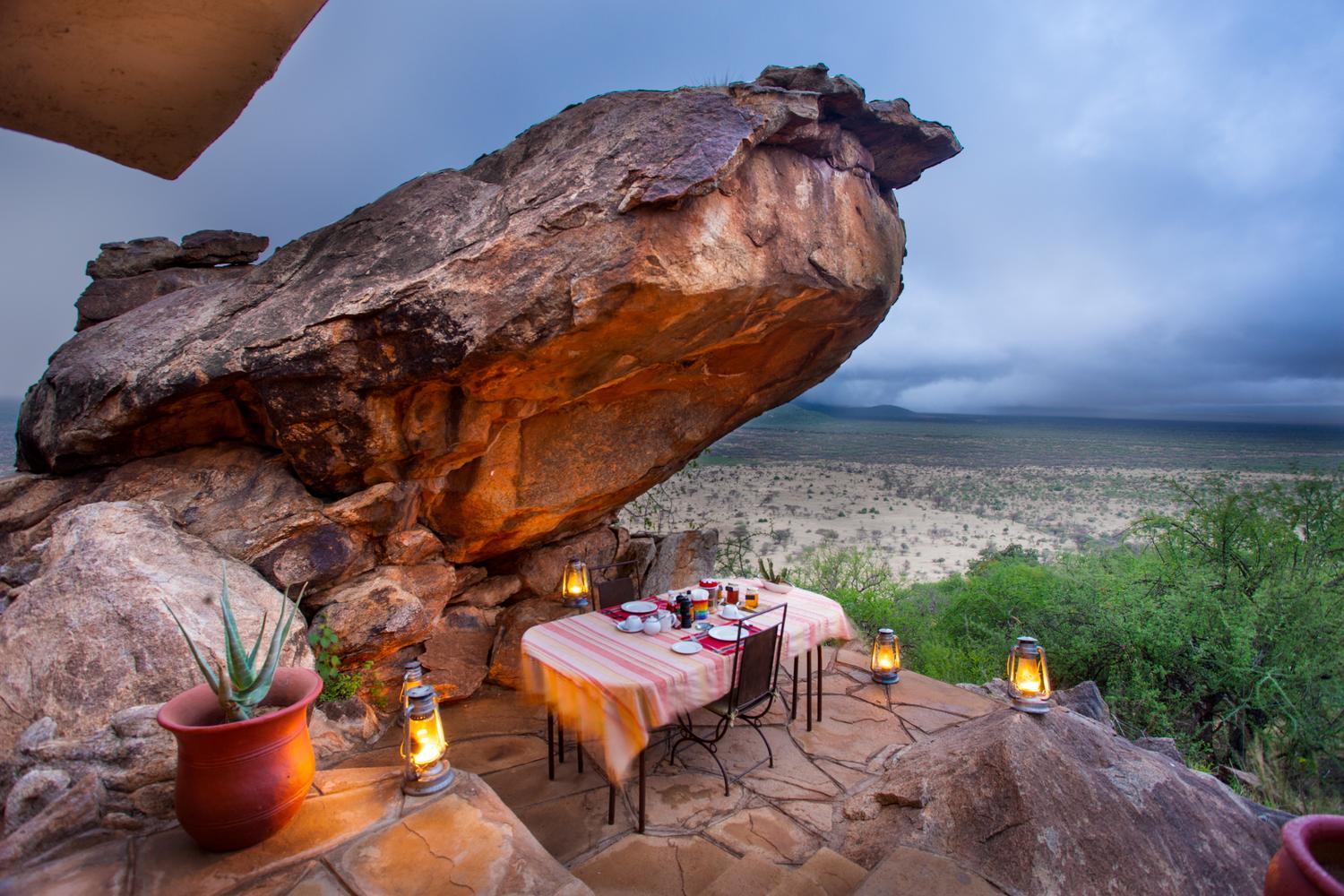 A breakfast table with lanterns set under a massive boulders overlooking a vast African savanna at Saruni Samburu.