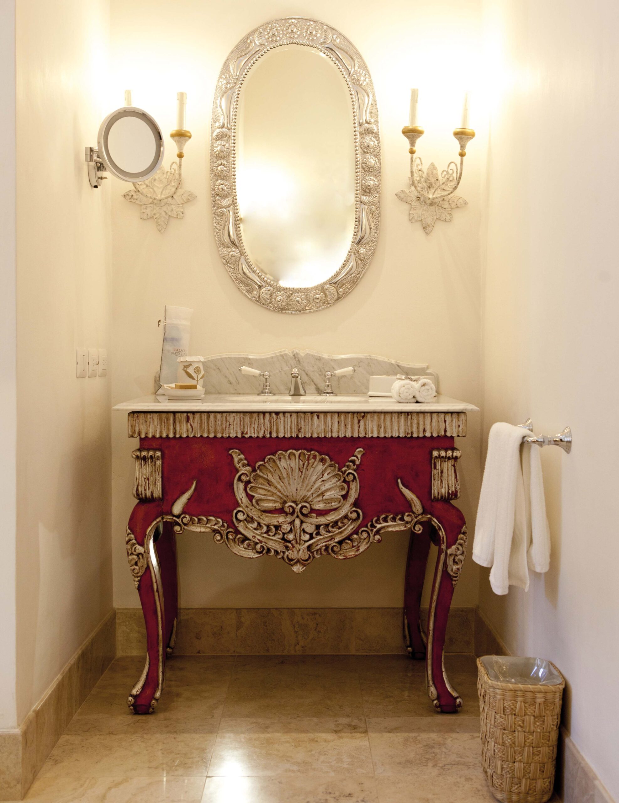Elegant bathroom with ornate mirror, red vintage vanity, and wall sconces.