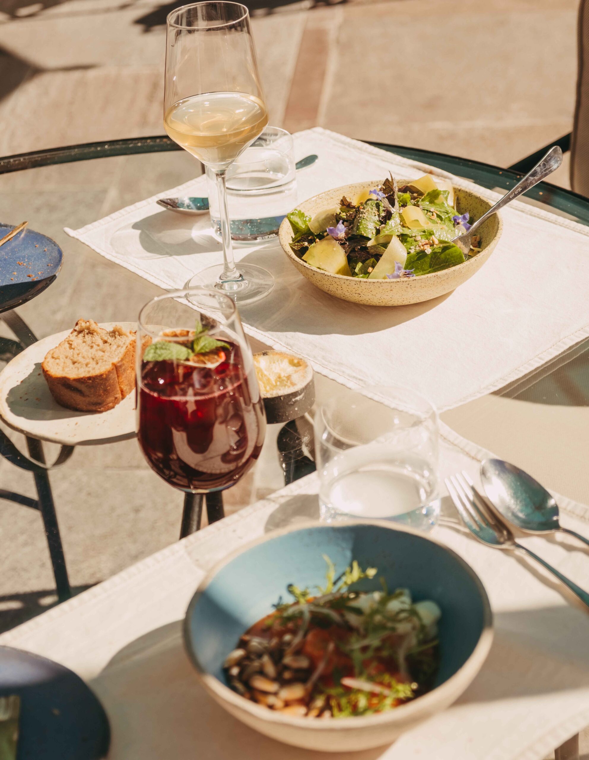 Outdoor meal with salads, bread, and glasses of wine on a sunny table.