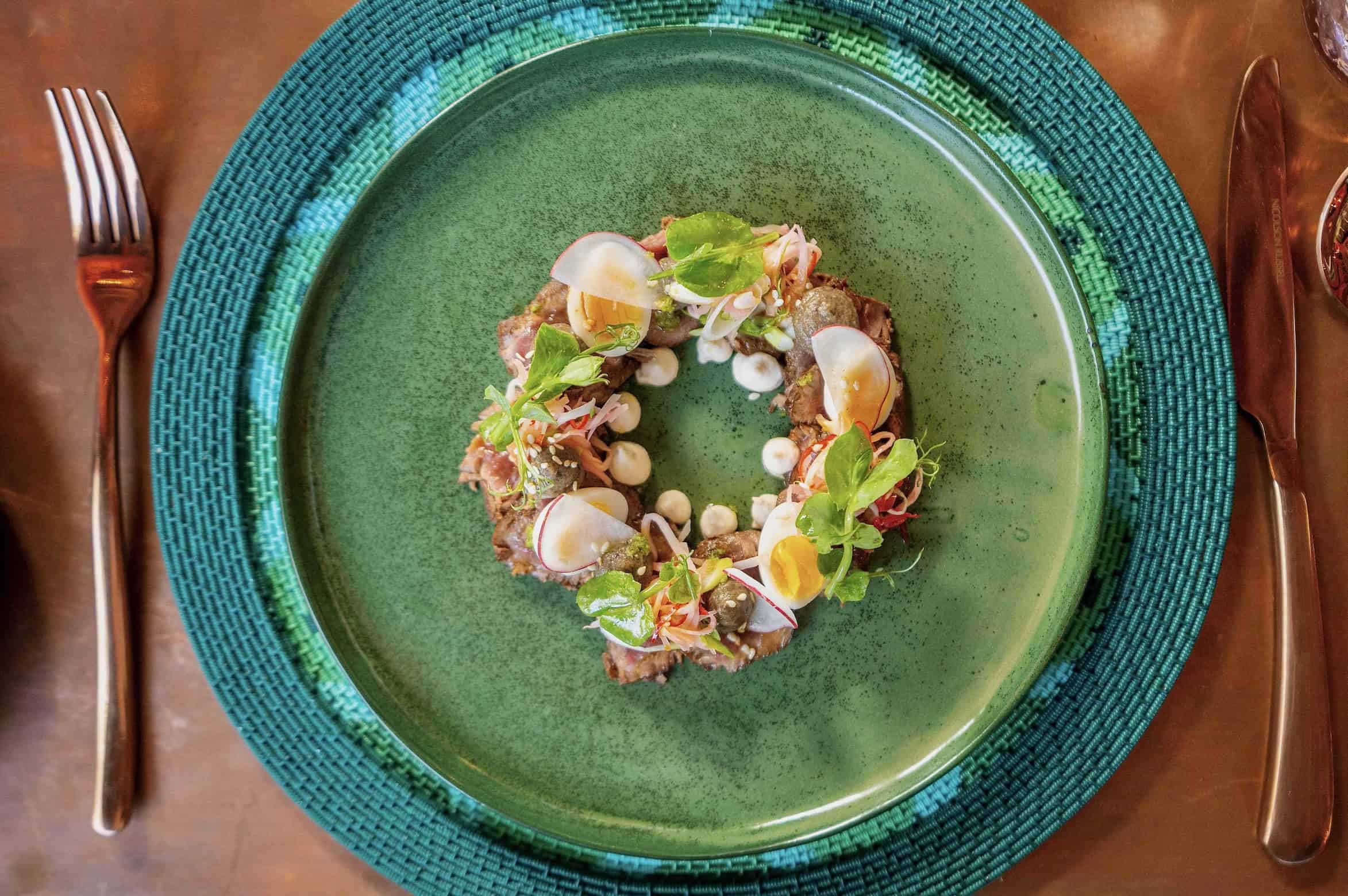 Aerial shot of food on a gren plate with a copper knife and fork set beside it. The food is arranged in a ring shape, and is a salad made with egg, thinly sliced radish and herbs