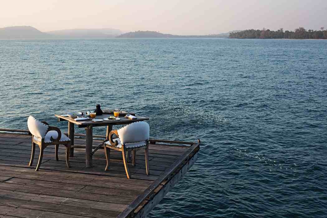 A wooden table and two white chairs on a deck over the ocean with distant hills in the background.