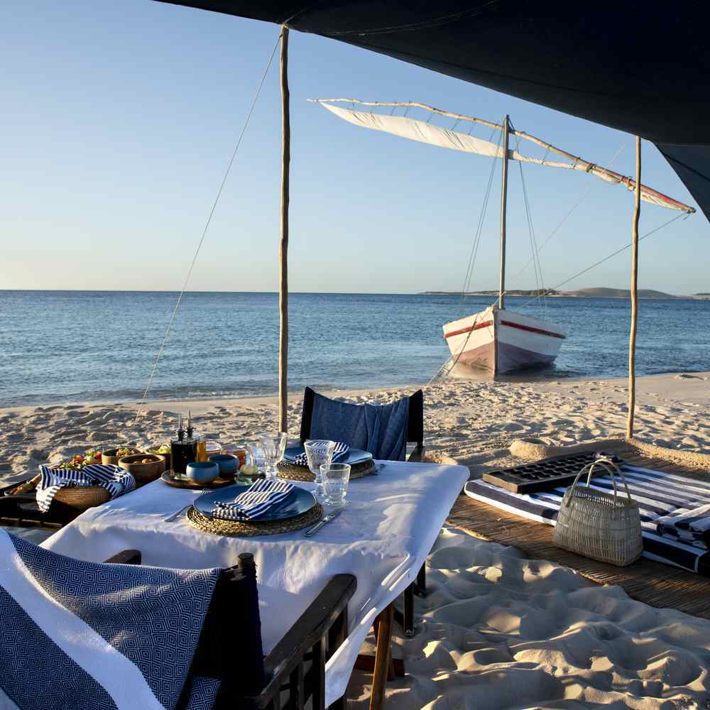 A picnic on a beach. The set up overlooks turquoise waters and a traditional boat.