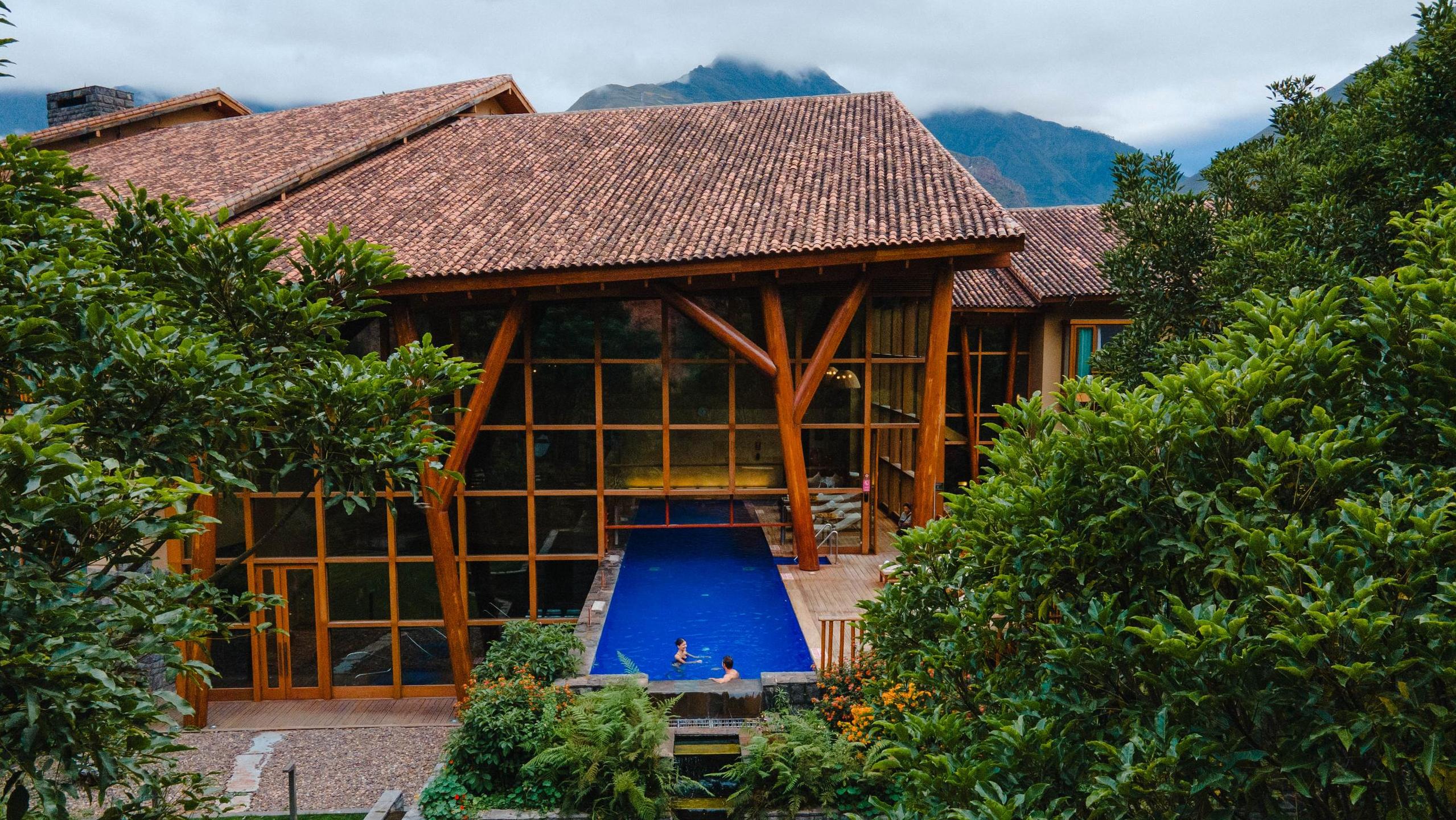 Wooden house with large windows, a blue pool, surrounded by lush greenery and mountains in the background.