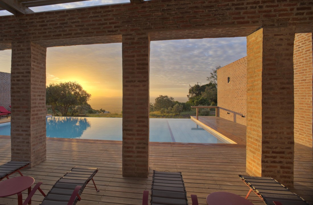 View through brick columns of a swimming pool at sunset with lounge chairs on a wooden deck.
