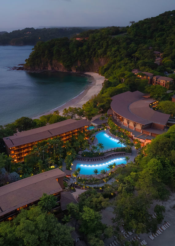 Aerial view of a luxury resort with glowing blue pools and coastline buildings at dusk.