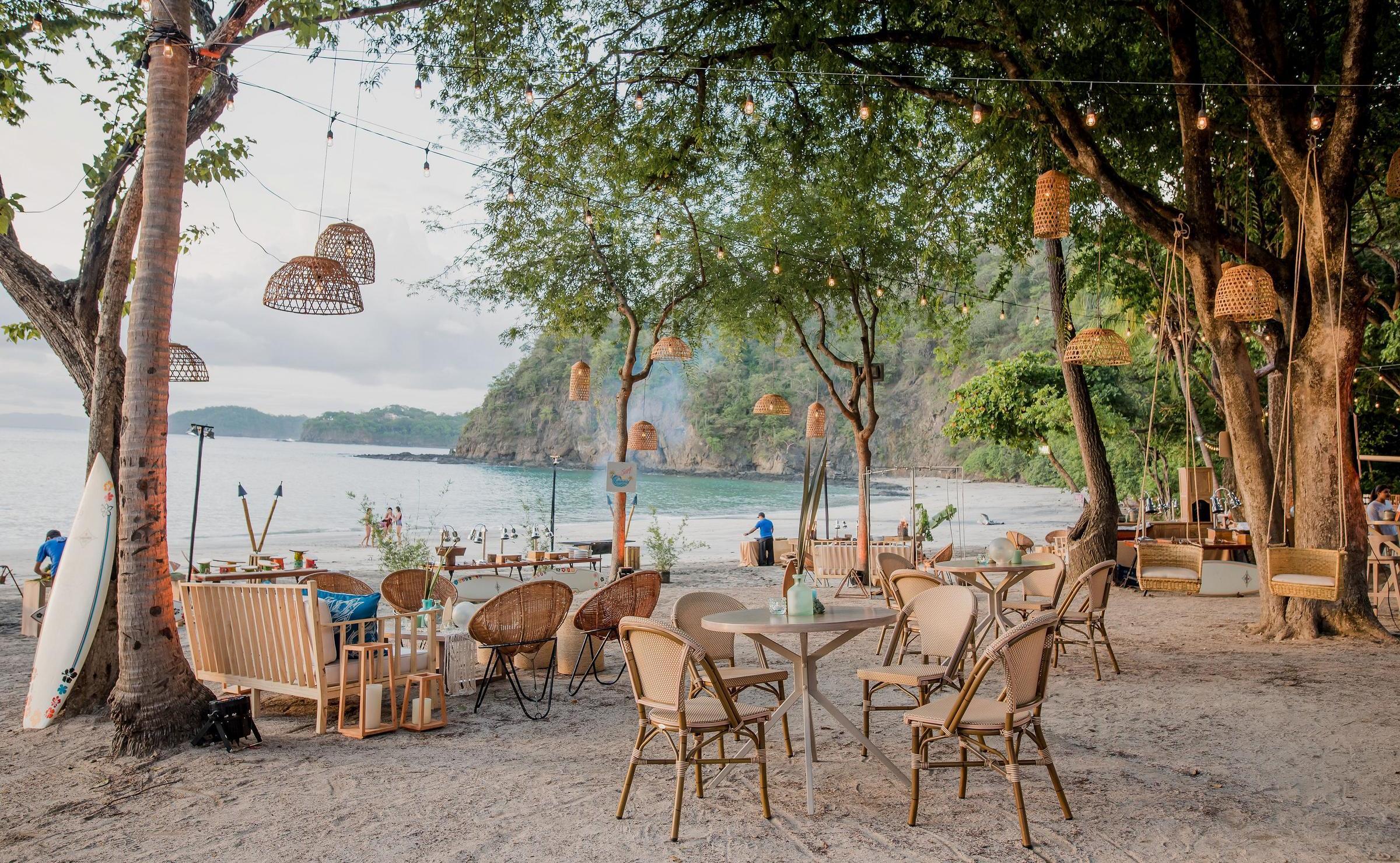 Beachside dining area with hanging woven lamps, wooden furniture, and a surfboard leaning against a tree.