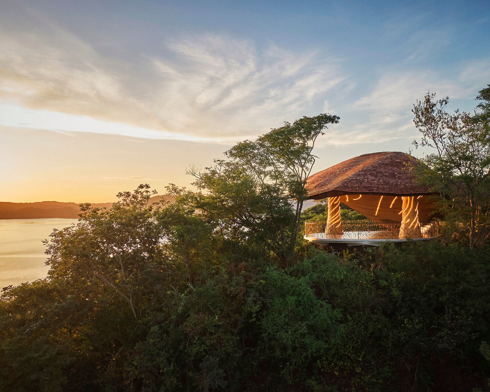 An illuminated circular wooden pavilion nestled in green trees on a cliffside at sunset.