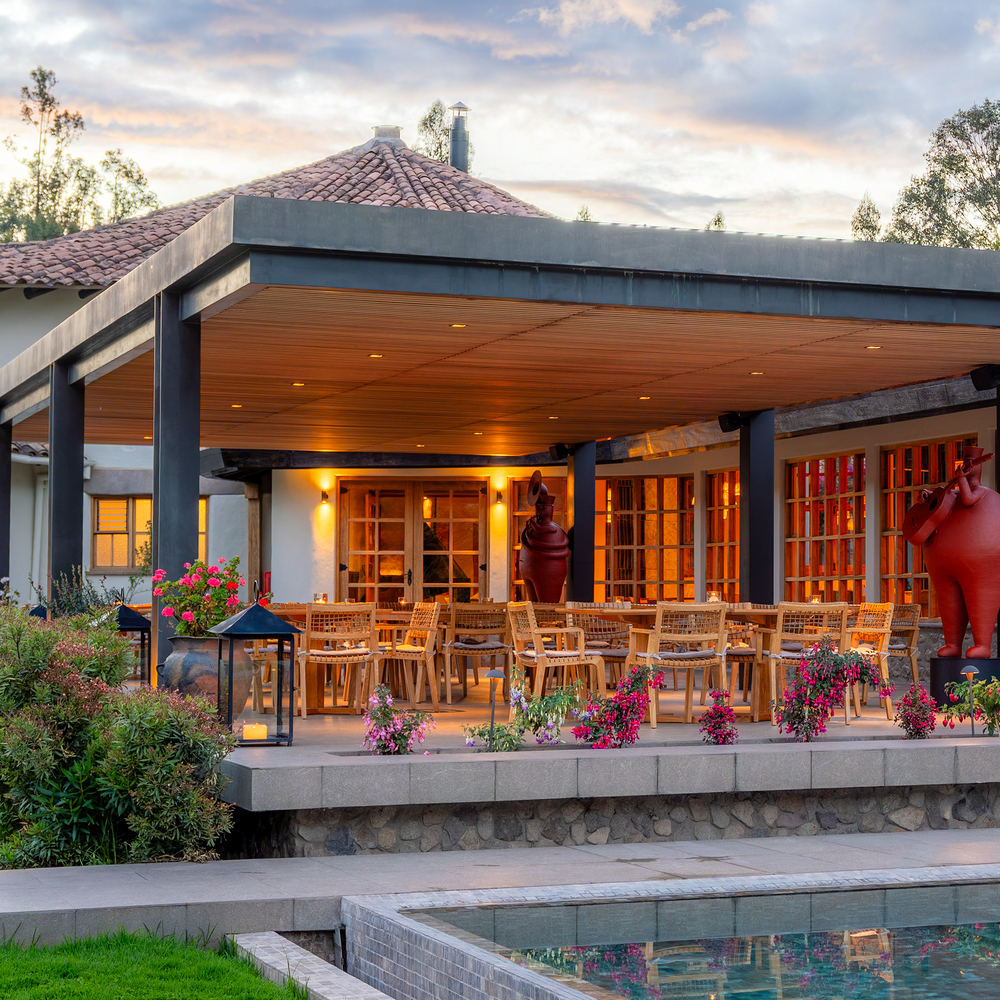 Covered outdoor dining area with a long wooden table, woven chairs, and pink flowers near a pool.