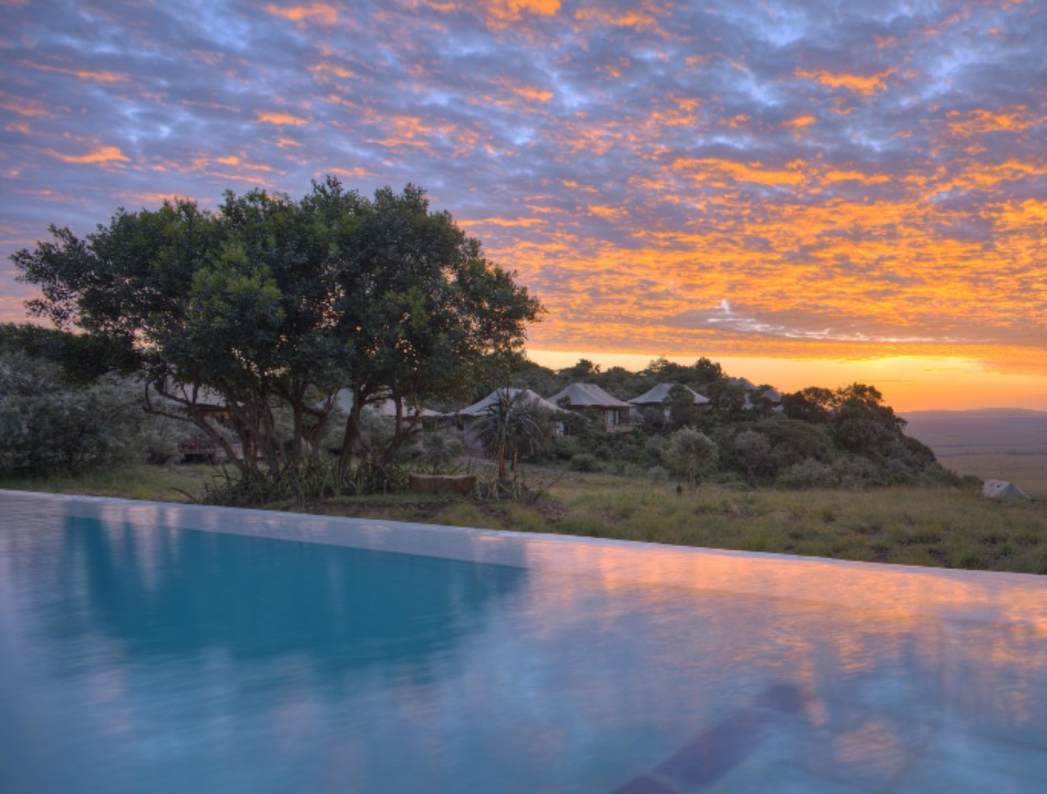 A calm infinity pool in the foreground reflects a vibrant orange sunset sky with safari tents and trees behind it.