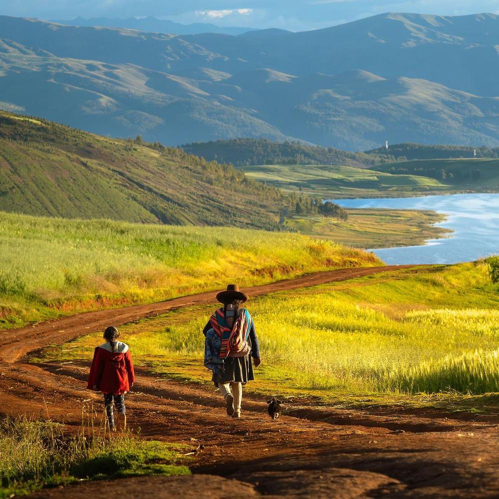 A person and a child in a red coat walk on a dirt path toward a lake surrounded by green hills.