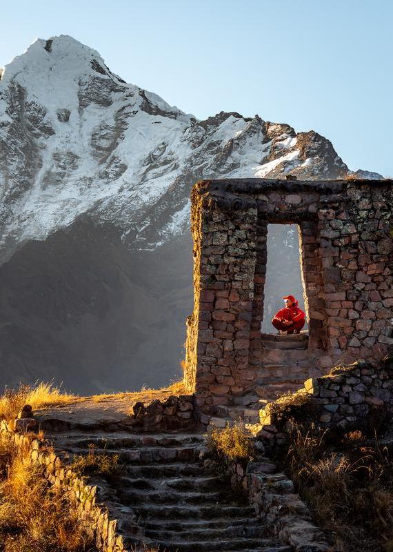 A person in a red jacket sitting inside a stone ruin window, with stone steps and a snowy mountain.