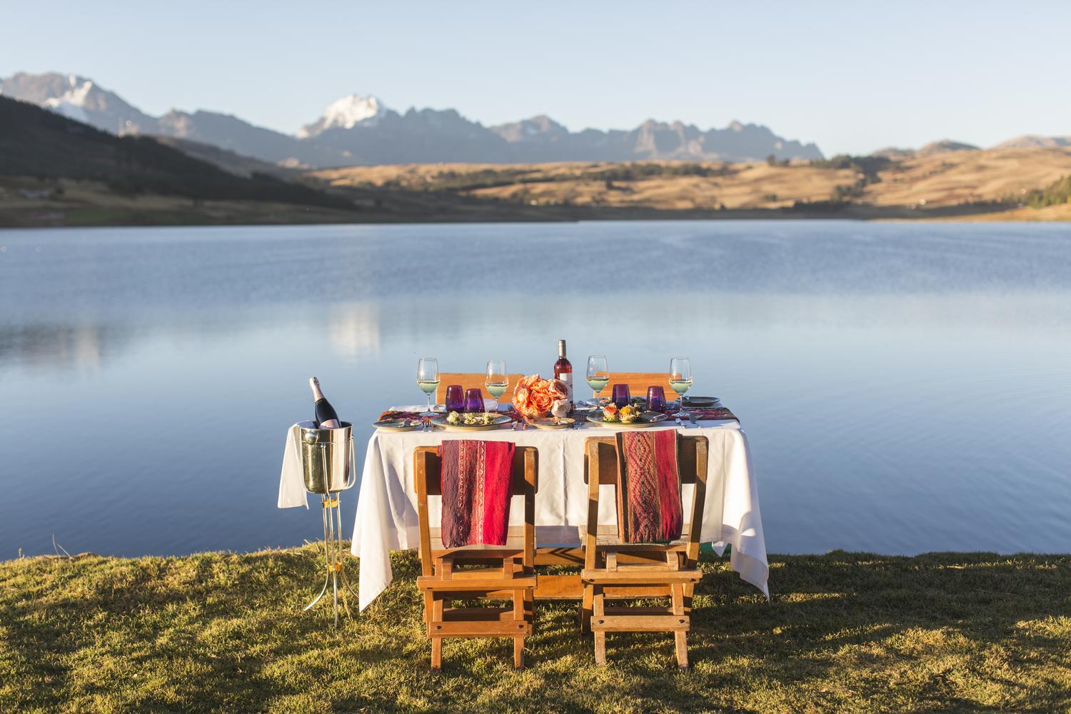A dining table set for two on a grassy lakeshore, with snow-capped mountains in the background.