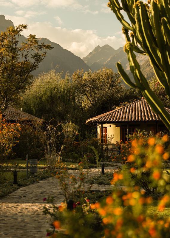 Stone pathway leading to a yellow building with a tiled roof, framed by a cactus and mountains.
