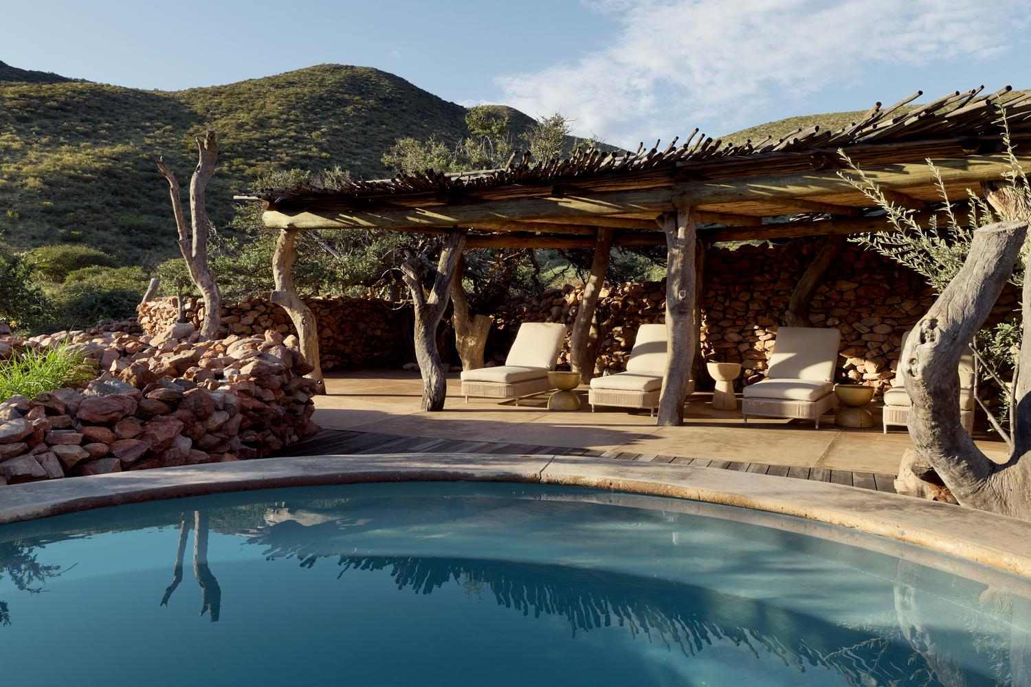 A curved swimming pool in front of a shaded wooden deck with comfortable sun loungers.