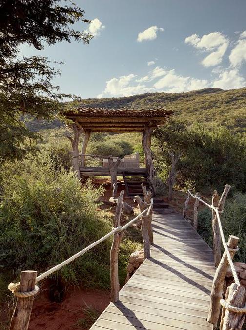 A wooden plank walkway with rope handrails leading to a thatched gazebo in a natural landscape.