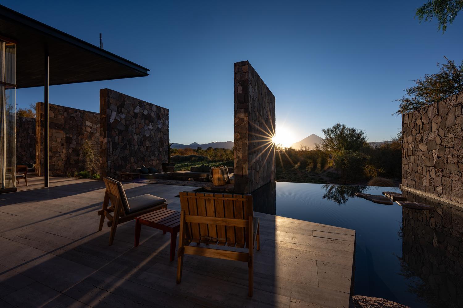 Sunburst appearing past stone privacy walls onto an outdoor patio with wooden chairs next to a calm reflecting pool.