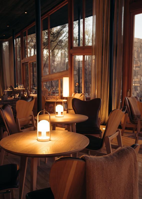 Interior view of a hotel lounge featuring round wooden tables illuminated by glowing dome lamps and cozy chairs.