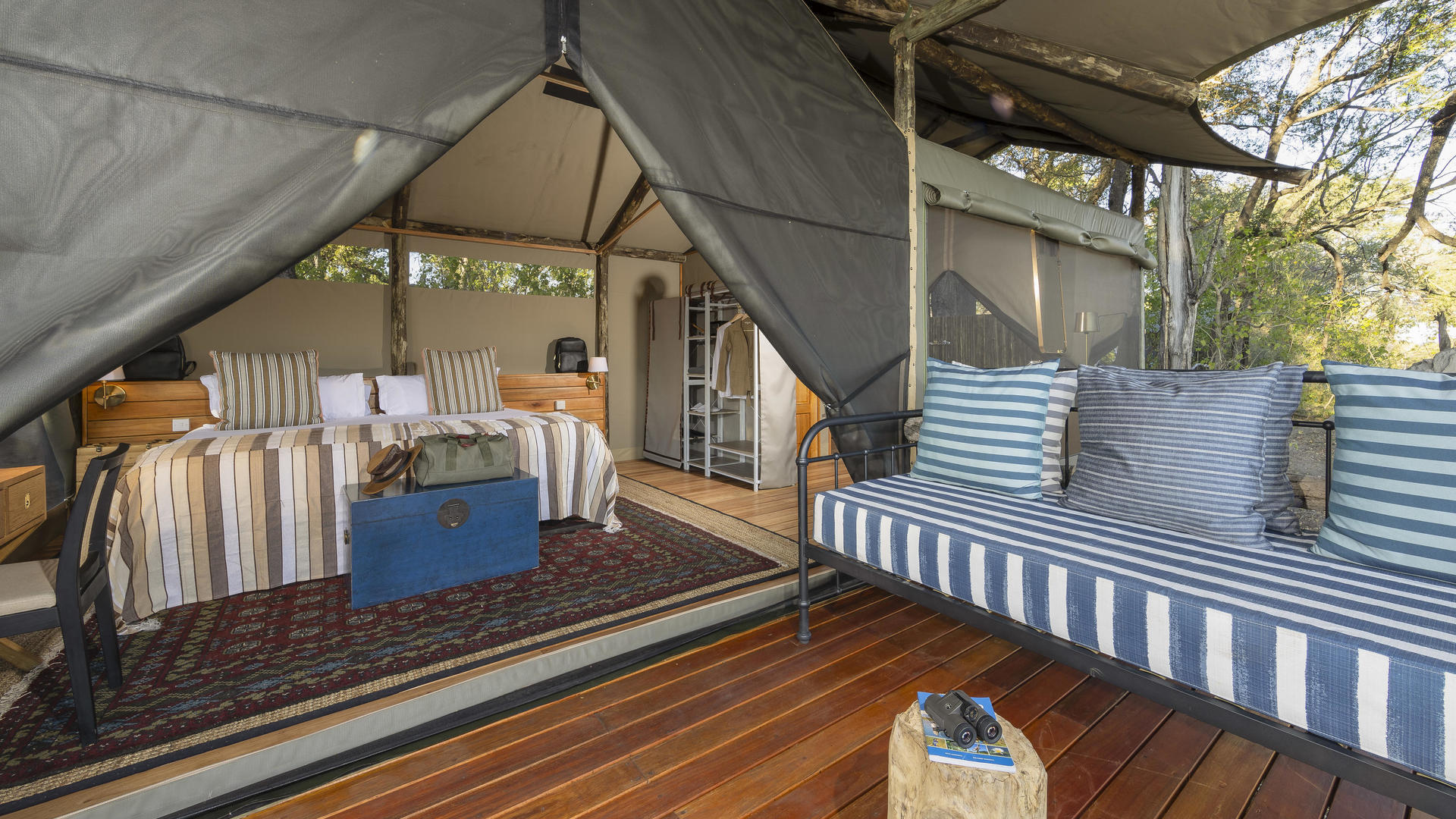 Inside a safari tent showing a bed with striped pillows, a blue trunk, and a striped outdoor sofa.