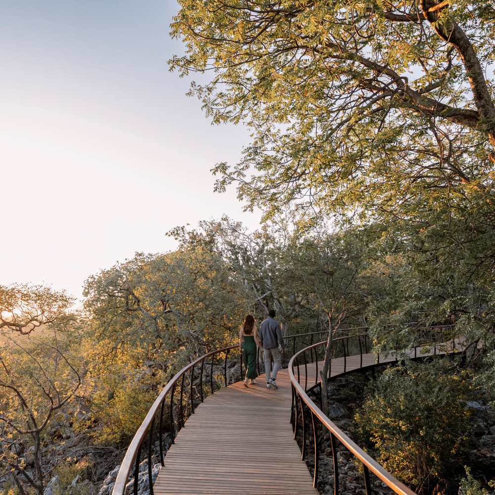 A couple walks hand-in-hand along a winding wooden walkway surrounded by green trees at sunset.